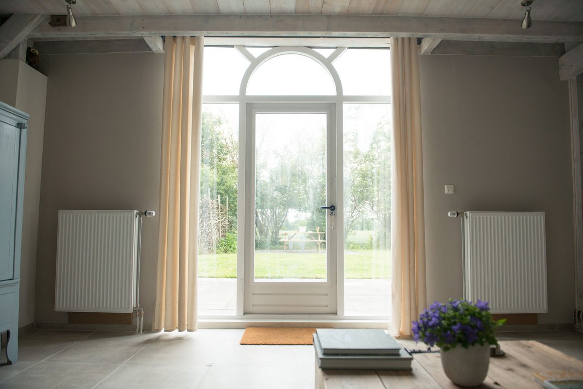 Salón con gran ventana en arco, cortinas al suelo y vistas al jardín – Zeeuwshuisje casa de vacaciones, Koudekerke